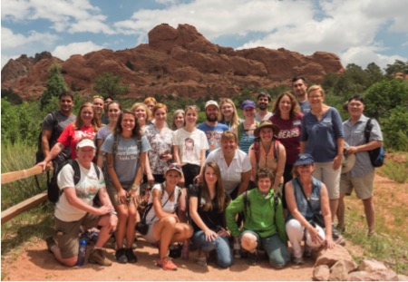 A photo of past mentors during an in-person gathering in Colorado in 2016.  About twenty young adults are standing outdoors on a sunny day with a large rock formation in the background.